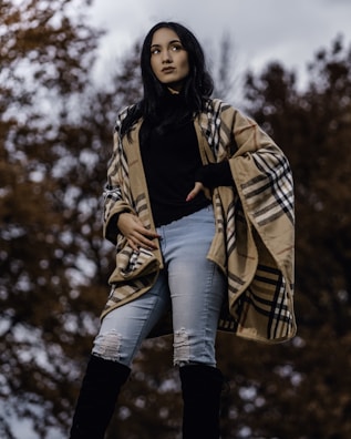 A person standing outdoors wearing a stylish plaid poncho, black top, light blue ripped jeans, and knee-high black boots. The background features trees with autumn leaves and an overcast sky, creating a moody atmosphere.