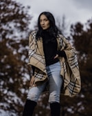 A person stands outdoors wearing a plaid shawl, black top, ripped jeans, and knee-high boots. The background consists of blurred autumnal trees.