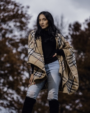 A person stands outdoors wearing a plaid shawl, black top, ripped jeans, and knee-high boots. The background consists of blurred autumnal trees.