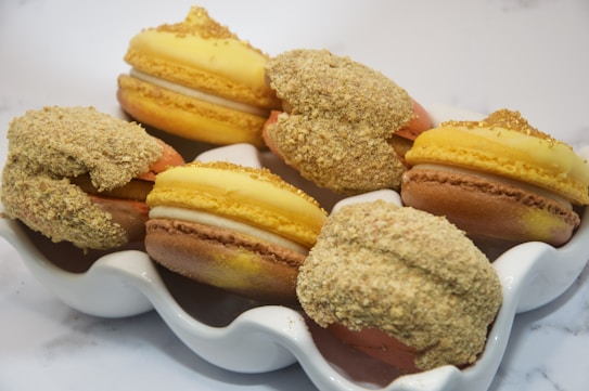 A decorative plate holds a selection of macarons with vanilla and golden hues. Each macaron is topped with a crumbly texture, possibly a nut or cookie topping, and has a smooth cream filling in between the shells. The background is minimalist, highlighting the dessert's intricate details.