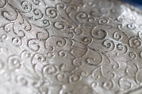 An intricate fabric pattern featuring swirling designs on a shiny, textured surface. The material has a metallic sheen, adding a luxurious and elegant appeal.