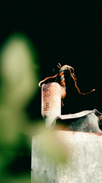 A rusty, old metal tool with a spiral of wires on top, set against a blurred dark green background. The texture of the rust and metal surface is prominent, creating a sense of age and wear.