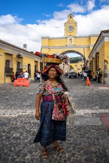 A person stands on a cobblestone street wearing colorful traditional clothing, including a woven skirt, blouse, and textile accessories. They have a basket of textiles balanced on their head. The background features a historical yellow arch with a clock and a clear blue sky with some clouds. Several people are walking in the street behind.