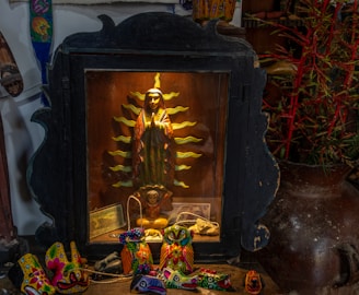 A display featuring a religious icon of the Virgin Mary surrounded by radiant golden rays, housed in a dark wooden frame. Below the icon are brightly-colored decorative objects with intricate patterns. These objects resemble small masks or figures with vivid hues of red, yellow, and green. To the right, some decorative sticks with red accents are visible.