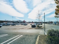 A large parking lot filled with numerous cars is visible. The lot is adjacent to a row of stores with recognizable signs, including officesupply and household goods stores. The sky is mostly clear with a few clouds, and the sunlight casts shadows on the ground. Sparse trees with few leaves are also present in the scene.