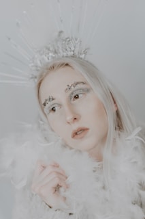 A person with long, light-colored hair is adorned with an intricate silver headpiece resembling a crown. Their makeup is elaborate, featuring rhinestones above the eyebrows, and they wear a fluffy, light-colored garment that seems to be made of soft feathers or faux fur. The overall color palette is soft and muted, lending a dreamy or ethereal quality to the scene.