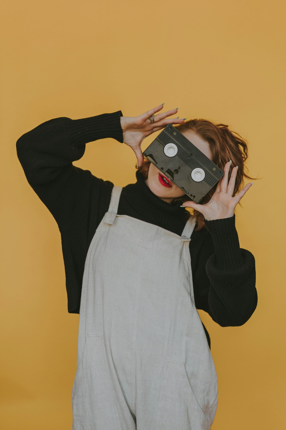 a woman wearing overalls and a black sweater is holding a camera up to her