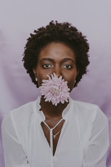 A person wearing a white blouse holds a pink flower in front of their face, with a soft purple background. Their facial expression appears calm and serene.