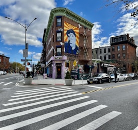 A street corner with a vibrant mural on the side of a brick building. The mural features a prominent figure with an orange and yellow sunburst background. The building has a unique architectural design with a green cornice. There are several parked cars along the street, and a well-marked pedestrian crossing. People are visible walking on the sidewalk, and the sky is partly cloudy.