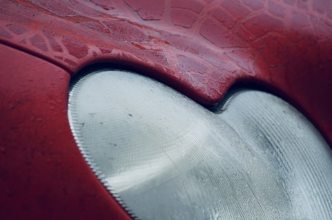 Close-up view of a car's headlight with a red body exterior. The texture on the red paint is rough and resembles a web-like pattern. The headlight appears to have a frosty or matte finish.