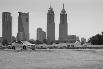 A modern cityscape featuring tall skyscrapers with unique architectural designs in the background. A sleek, white car is parked on a sandy foreground, with trees and urban buildings surrounding the area.