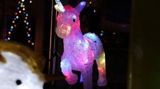 A decorative unicorn sculpture illuminated with vibrant multicolored lights. The unicorn has a translucent appearance, allowing the lights to create a glowing effect, especially in shades of pink, blue, and yellow. In the background, strings of small, colorful lights create a festive atmosphere.