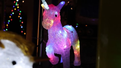A decorative unicorn sculpture illuminated with vibrant multicolored lights. The unicorn has a translucent appearance, allowing the lights to create a glowing effect, especially in shades of pink, blue, and yellow. In the background, strings of small, colorful lights create a festive atmosphere.