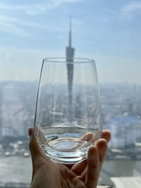A clear glass is held up by a hand against a background of a city skyline with a tall, distinctive tower. The focus is primarily on the glass, while the skyline and tower appear blurred, creating a depth effect.