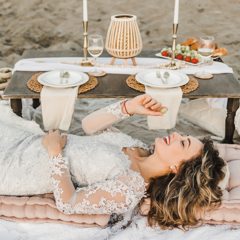 A woman in a lace dress is lying on a cushioned area near a beach, smiling and holding a grape. In front of her is a low wooden table set for two with elegant place settings, wine glasses, and a variety of foods including fruits and pastries. Decorative elements such as candles and a woven lamp are also present, creating a romantic and serene atmosphere.