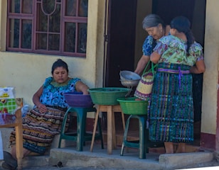 Three women are gathered outside a building, dressed in colorful traditional attire. One woman is seated, reaching into a basket on a plastic stool, while the other two are standing, examining a metal bowl and a woven basket. The setting appears to be a casual outdoor market or a home front.