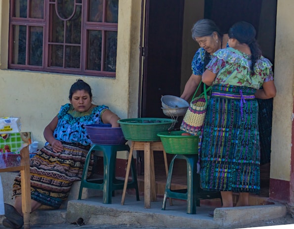Three women are gathered outside a building, dressed in colorful traditional attire. One woman is seated, reaching into a basket on a plastic stool, while the other two are standing, examining a metal bowl and a woven basket. The setting appears to be a casual outdoor market or a home front.