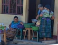 Three women are gathered outside a building, dressed in colorful traditional attire. One woman is seated, reaching into a basket on a plastic stool, while the other two are standing, examining a metal bowl and a woven basket. The setting appears to be a casual outdoor market or a home front.