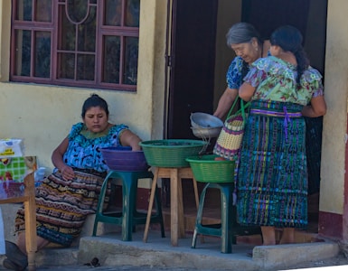 Three women are gathered outside a building, dressed in colorful traditional attire. One woman is seated, reaching into a basket on a plastic stool, while the other two are standing, examining a metal bowl and a woven basket. The setting appears to be a casual outdoor market or a home front.