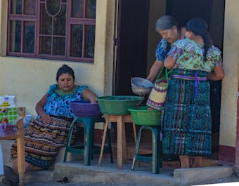 Three women are gathered outside a building, dressed in colorful traditional attire. One woman is seated, reaching into a basket on a plastic stool, while the other two are standing, examining a metal bowl and a woven basket. The setting appears to be a casual outdoor market or a home front.