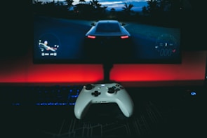 A gaming setup featuring a white controller in the foreground, with a racing video game displayed on a large monitor. The background is illuminated with a red light, creating a dramatic contrast with the dark surroundings. A keyboard and mouse are also visible.