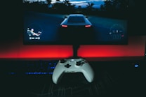 A gaming setup featuring a white controller in the foreground, with a racing video game displayed on a large monitor. The background is illuminated with a red light, creating a dramatic contrast with the dark surroundings. A keyboard and mouse are also visible.