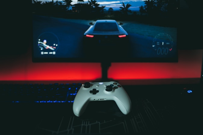 A gaming setup featuring a white controller in the foreground, with a racing video game displayed on a large monitor. The background is illuminated with a red light, creating a dramatic contrast with the dark surroundings. A keyboard and mouse are also visible.