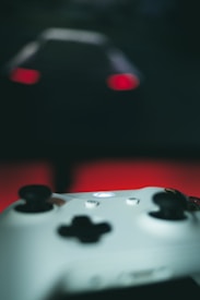 A close-up of a white gaming controller with dark buttons and a glowing central button. In the blurred background, red lights suggest the taillights of a vehicle against a dark backdrop.