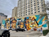 A vibrant mural covers the side of a building, depicting several faces wearing various headgear, including helmets and goggles. The artwork utilizes bold colors such as blues, oranges, and yellows, creating a dynamic and energetic composition. The mural is surrounded by a parking lot with several parked vehicles, and nearby buildings are visible, including one labeled 'Westin'.