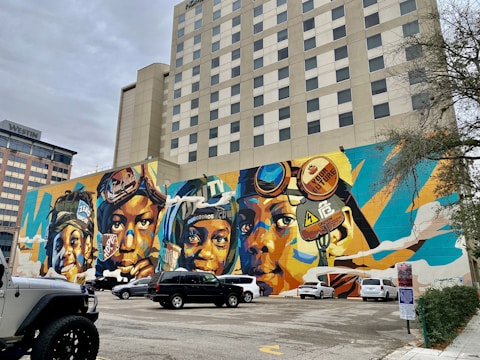 A vibrant mural covers the side of a building, depicting several faces wearing various headgear, including helmets and goggles. The artwork utilizes bold colors such as blues, oranges, and yellows, creating a dynamic and energetic composition. The mural is surrounded by a parking lot with several parked vehicles, and nearby buildings are visible, including one labeled 'Westin'.