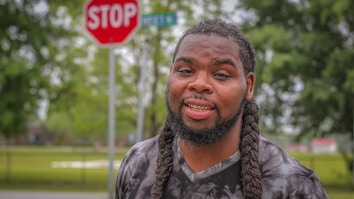 A man with braided hair and a gold grill is standing outdoors. Behind him, a stop sign and some greenery are visible, indicating a street setting. The man's expression is relaxed and he seems to be smiling slightly.