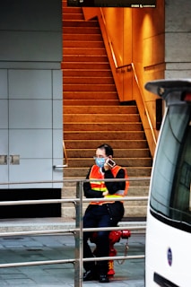 A person wearing an orange safety vest and a blue face mask, sitting on a red fire hydrant, talking on a cell phone. The background features a staircase with warm lighting and a building wall with a grid design. A railing is visible in the foreground, and part of a vehicle is on the right side of the image.
