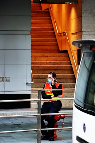 A person wearing an orange safety vest and a blue face mask, sitting on a red fire hydrant, talking on a cell phone. The background features a staircase with warm lighting and a building wall with a grid design. A railing is visible in the foreground, and part of a vehicle is on the right side of the image.