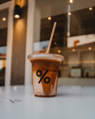 A cold beverage served in a clear plastic cup with a lid and a straw, featuring a bold black percentage symbol on the front. The drink consists of layers of a light brown coffee-like liquid over a paler, milkier substance. It is placed on a smooth white surface, with a blurred indoor background including lights and windows.