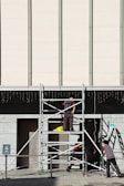 Three workers are building or inspecting scaffolding in front of a large modern building. The sun casts strong shadows, and the scene is set against a light-colored wall with vertical lines. A yellow plastic bag is seen on the scaffolding platform.