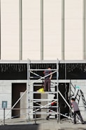 Three workers are building or inspecting scaffolding in front of a large modern building. The sun casts strong shadows, and the scene is set against a light-colored wall with vertical lines. A yellow plastic bag is seen on the scaffolding platform.