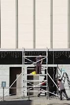 Three workers are building or inspecting scaffolding in front of a large modern building. The sun casts strong shadows, and the scene is set against a light-colored wall with vertical lines. A yellow plastic bag is seen on the scaffolding platform.