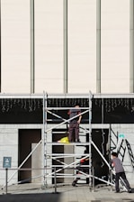 Three workers are building or inspecting scaffolding in front of a large modern building. The sun casts strong shadows, and the scene is set against a light-colored wall with vertical lines. A yellow plastic bag is seen on the scaffolding platform.