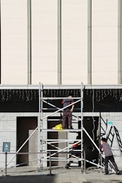 Three workers are building or inspecting scaffolding in front of a large modern building. The sun casts strong shadows, and the scene is set against a light-colored wall with vertical lines. A yellow plastic bag is seen on the scaffolding platform.