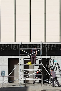 Three workers are building or inspecting scaffolding in front of a large modern building. The sun casts strong shadows, and the scene is set against a light-colored wall with vertical lines. A yellow plastic bag is seen on the scaffolding platform.