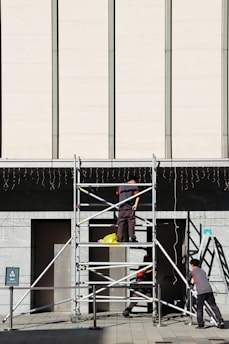 Three workers are building or inspecting scaffolding in front of a large modern building. The sun casts strong shadows, and the scene is set against a light-colored wall with vertical lines. A yellow plastic bag is seen on the scaffolding platform.