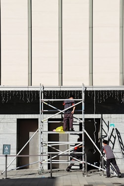 Three workers are building or inspecting scaffolding in front of a large modern building. The sun casts strong shadows, and the scene is set against a light-colored wall with vertical lines. A yellow plastic bag is seen on the scaffolding platform.