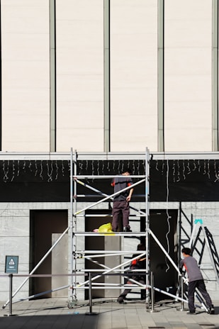 Three workers are building or inspecting scaffolding in front of a large modern building. The sun casts strong shadows, and the scene is set against a light-colored wall with vertical lines. A yellow plastic bag is seen on the scaffolding platform.