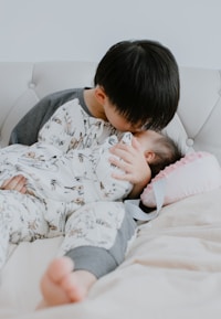 A young child lovingly kisses a baby on the forehead while they lie on a white tufted couch. Both children are dressed in matching pajamas with animal prints. The baby is swaddled and resting on a pink pillow with a ribbon.