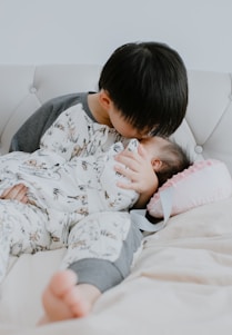 A young child lovingly kisses a baby on the forehead while they lie on a white tufted couch. Both children are dressed in matching pajamas with animal prints. The baby is swaddled and resting on a pink pillow with a ribbon.