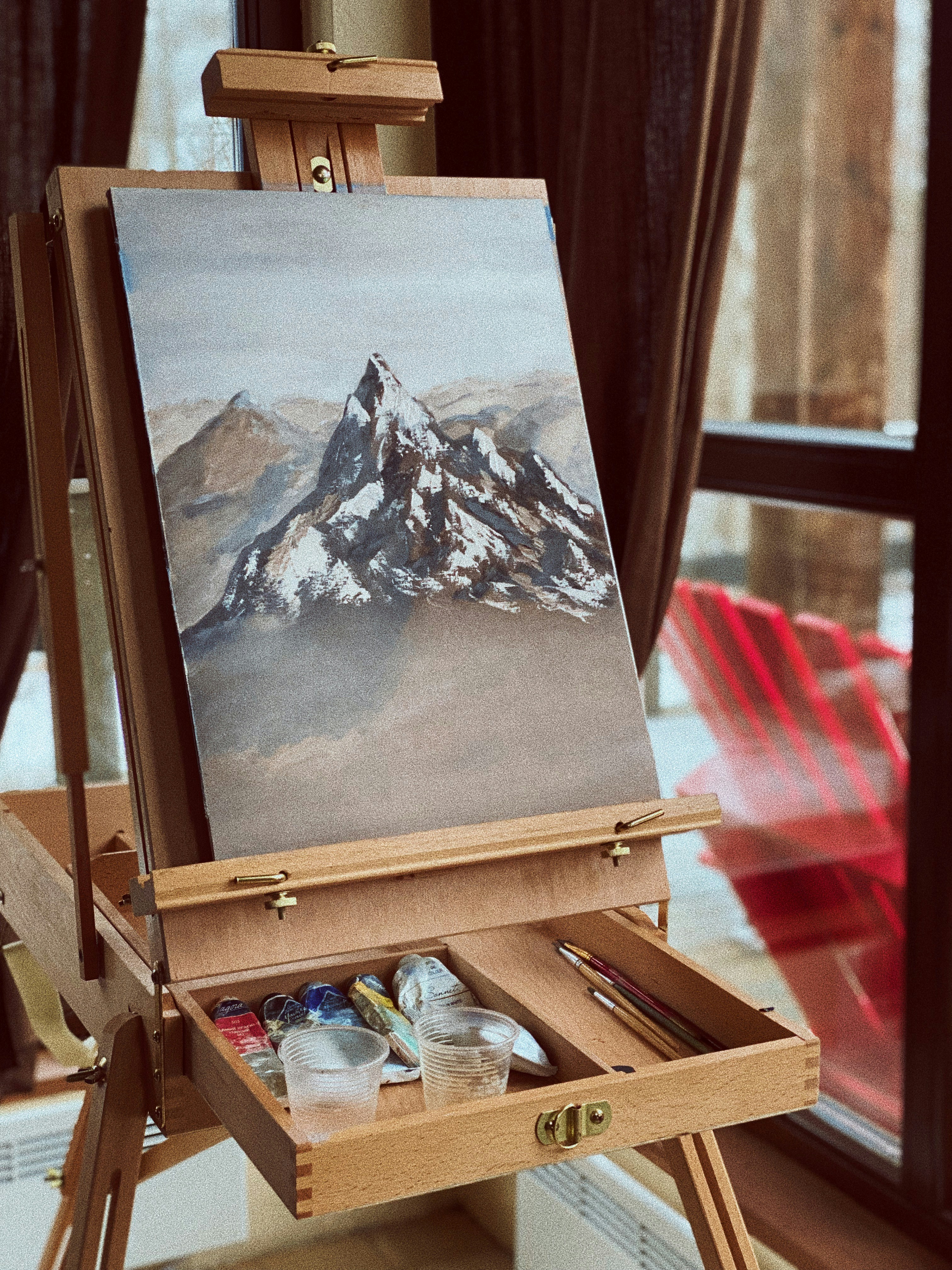 Mountain painting on a wooden box easel, paints, brushes and water cups in its open drawer