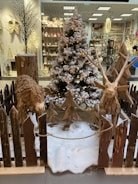 A festive display featuring a snow-covered Christmas tree adorned with lights stands in the center. Flanking the tree are two wooden sculptures of animals, crafted from logs. The scene is surrounded by a rustic wooden fence, with faux snow covering the ground. In the background, a store is visible, showcasing various holiday decorations and ornaments on shelves.