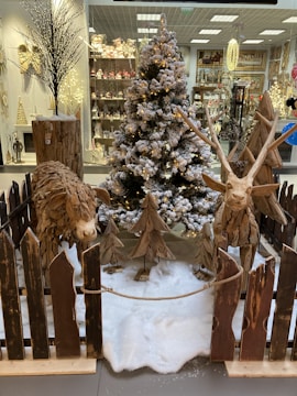A festive display featuring a snow-covered Christmas tree adorned with lights stands in the center. Flanking the tree are two wooden sculptures of animals, crafted from logs. The scene is surrounded by a rustic wooden fence, with faux snow covering the ground. In the background, a store is visible, showcasing various holiday decorations and ornaments on shelves.
