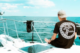 A person wearing a Versace collection t-shirt and a patterned cap is sitting on the deck of a boat. The individual faces the ocean, with calm blue water stretching to the horizon under a partly cloudy sky. A neatly coiled rope is secured to the railing nearby, and parts of the boat, including a railing and a winch, are visible.