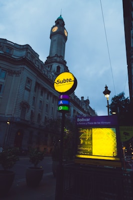 A street scene featuring a prominent building with a clock tower, characterized by its classic architecture. In the foreground, there is a brightly lit sign for the Buenos Aires subway system, labeled 'Subte', above station entrance labeled 'Bol&iacute;var'. The scene is captured at dusk or dawn, with street lamps providing additional lighting.